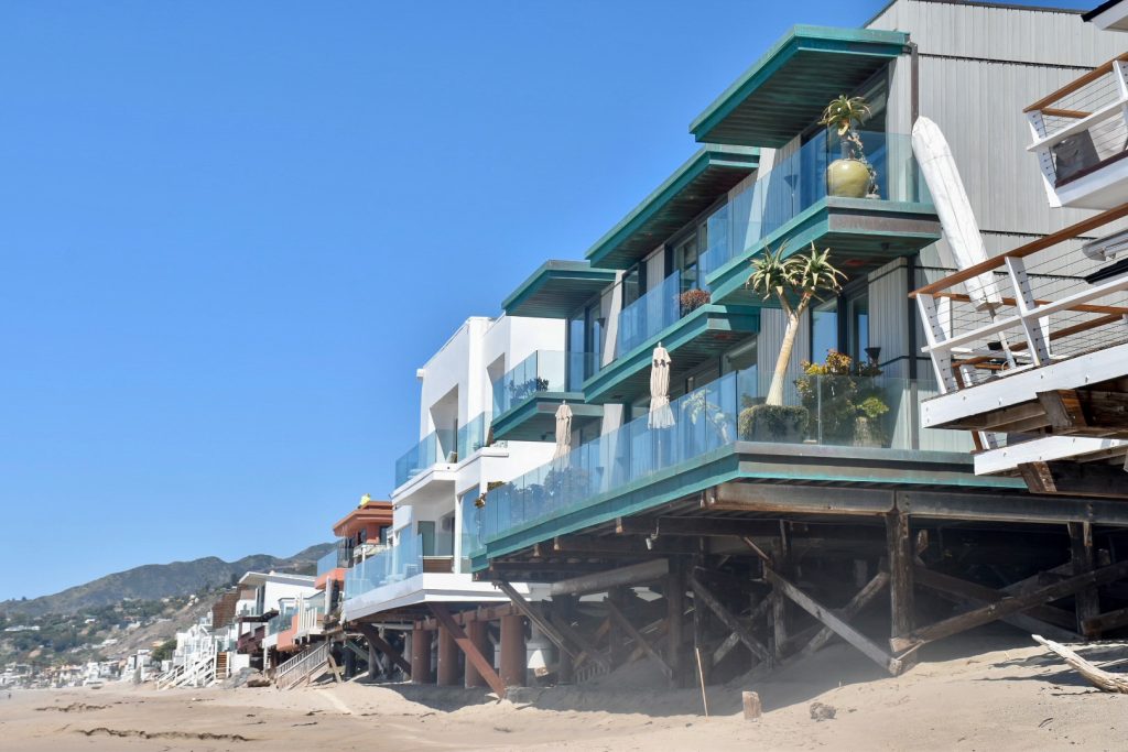 Sandy beach along the bottom. Clear blue sky to the left and top of photo. A row of impressive modern houses runs from the right hand side to the the distance on the left. The nearest house to the right hand side has green awnings and glass balcony walls and potted plants as decoration