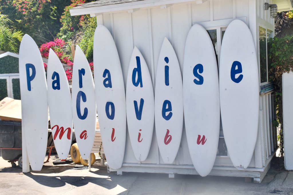 Row of white surf boards comprising a sign advertising 'Paradise Cove Malibu'. The sign is attached to a white hut and there are gardens behind