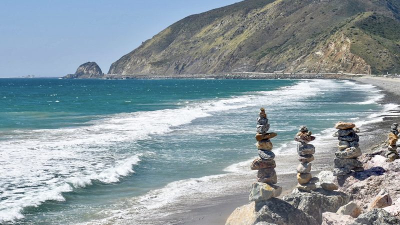 A road sweeps around a curved bay on the right hand side, green hills behind and pale blue sky above. The water in the bay is turquoise with white tipped waves coming to shore. Piles of stones sit in the foreground to the left