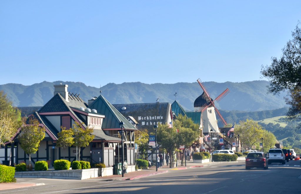 Scene of a High Street with row of shops to the left and a road with cars to the right. Hills behind and pale blue sky at the top. There is a windmill with red sails to the right of the High Street and one of the shops features the wording 'Birkholm's Bakery'