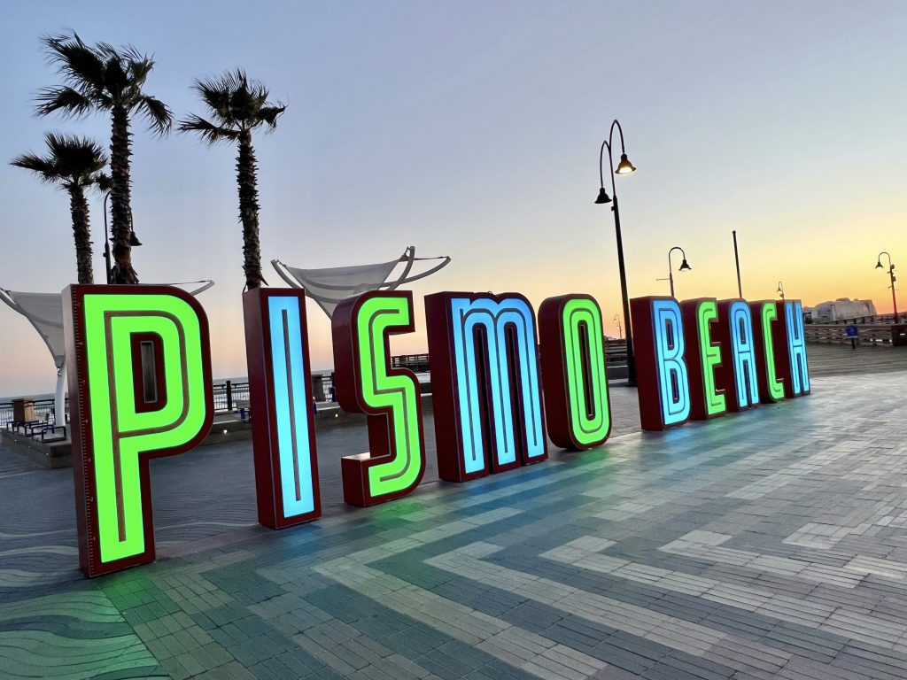 Sunset photo of a beach town sign which is illuminated in green and turquoise letters spelling 'PISMO BEACH'. Three palm trees stand behind to the left. Lamps stand behind to the right and middle right photo shows the orange glow of sunset. Striped pavement tiles in front