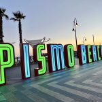 Sunset photo of a beach town sign which is illuminated in green and turquoise letters spelling 'PISMO BEACH'. Three palm trees stand behind to the left. Lamps stand behind to the right and middle right photo shows the orange glow of sunset. Striped pavement tiles in front