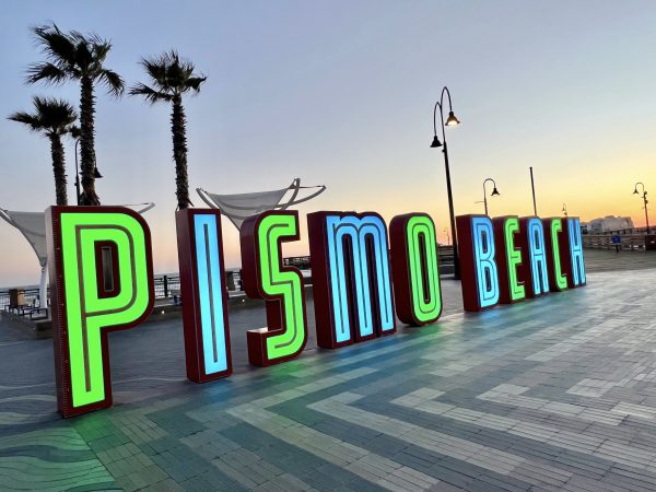 Sunset photo of a beach town sign which is illuminated in green and turquoise letters spelling 'PISMO BEACH'. Three palm trees stand behind to the left. Lamps stand behind to the right and middle right photo shows the orange glow of sunset. Striped pavement tiles in front