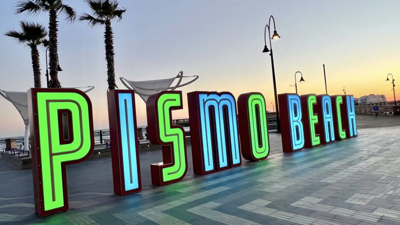 Sunset photo of a beach town sign which is illuminated in green and turquoise letters spelling 'PISMO BEACH'. Three palm trees stand behind to the left. Lamps stand behind to the right and middle right photo shows the orange glow of sunset. Striped pavement tiles in front