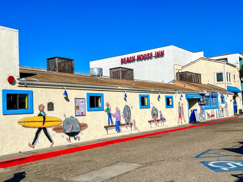 Bright blue sky above a long cream and white building across the middle of the photo - features a number of painted figures including a surfer carrying a board, along the building. Road in the bottom right of the photo