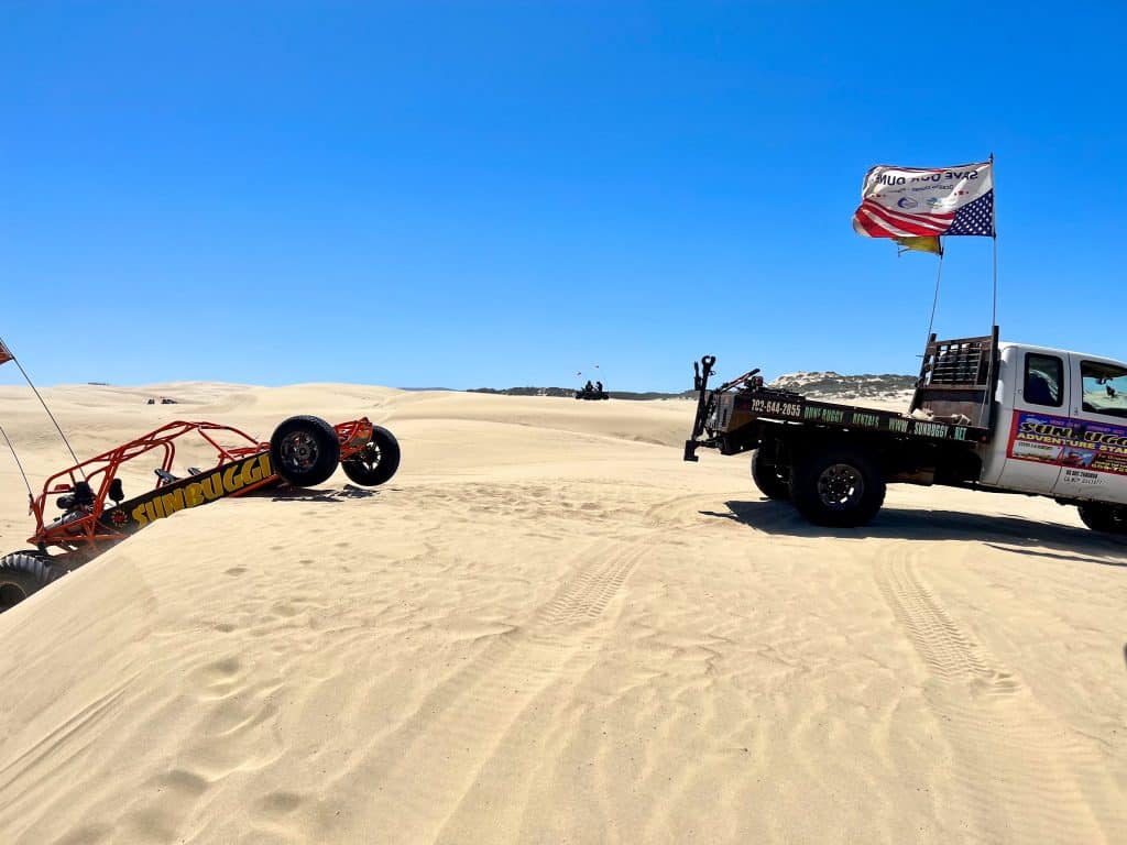 Bottom half features large expanse of sand. Top half is deep blue clear sky. A van with white cabin and two flags flying from the back is towing an orange, long wheel based buggy out of a sand dune - the buggy is on the left hand side