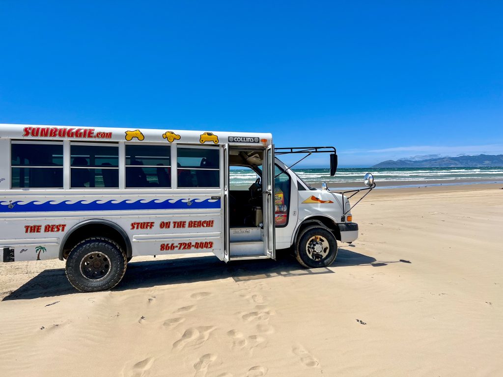 Flat golden sand at the bottom. Clear blue sky above. On the horizon is a strip of blue-green sea with white waves and some hills in the distance. A white school style bus is parked on the sand in the middle left. It says SunBiggie.com and 'The best stuff on the beach' with a phone number. It's door is open, no passengers visible