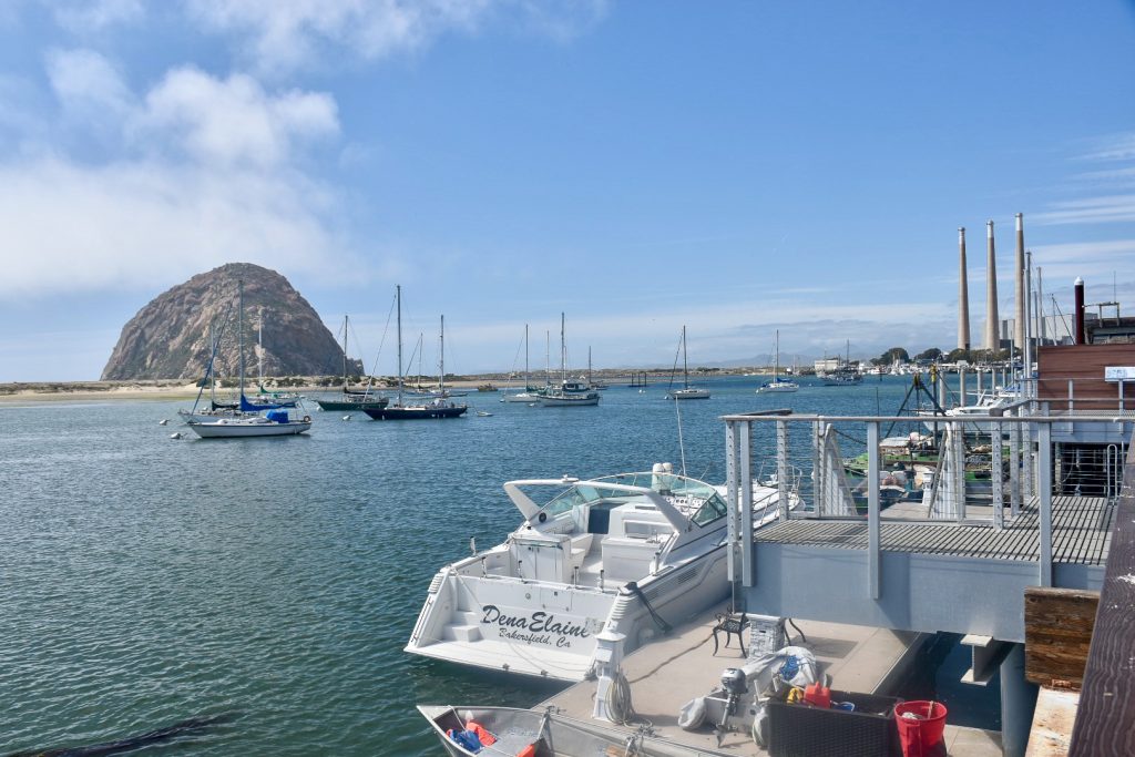 Bottom half of photo is blue body of water with sailboats parked up in the water. Buildings to the right hand side with power boat parked outside. across the horizon is a sandy beach and features a large circular rock to the left hand side. Blue sky above