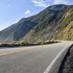 A wide grey road with double yellow lines in the middle, starts in the bottom left of the road and heads to the middle of the frame before winding round to the left along the cost of some rocky cliffs. Blue sky above and the sign of blue sea to the left of the road