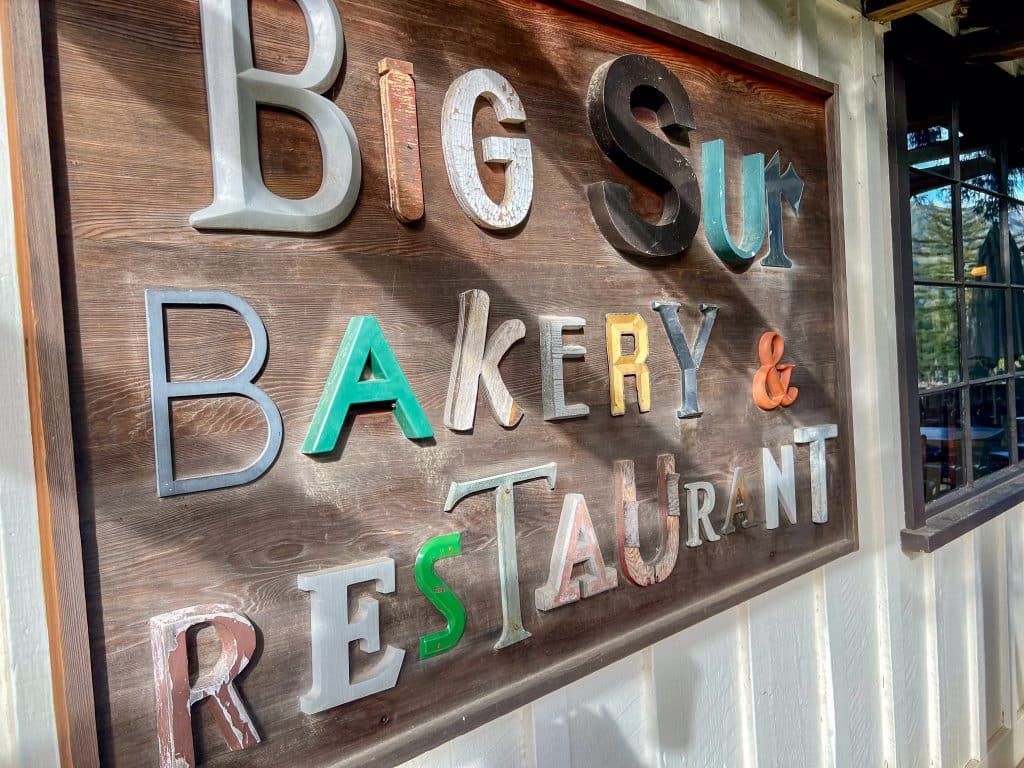 Brown wooden sign on the side of a wooden building painted cream. The sign says 'Big Sur Bakery and Restaurant' in multicoloured and sized letters. A window sits to the right of the sign