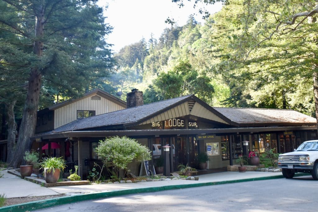 A single floor building, with a sign 'Big Lodge Sur' over the door surrounded by trees. A white van parked in front of the building. Sky is white and bleached out