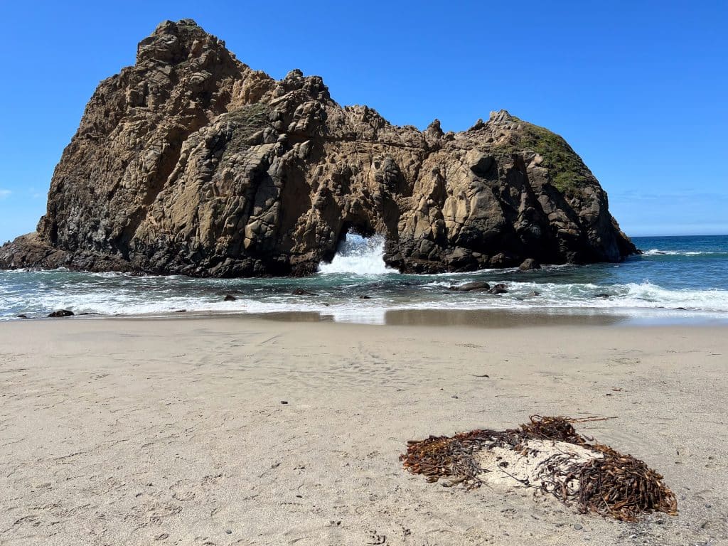 Golden sand with a small patch of seaweed in the bottom,. Clear blue sky above. Strip of blue sea across the middle, and large rock formation rising from the waves. It has a key hole in the middle through which waves are crashing towards the camera