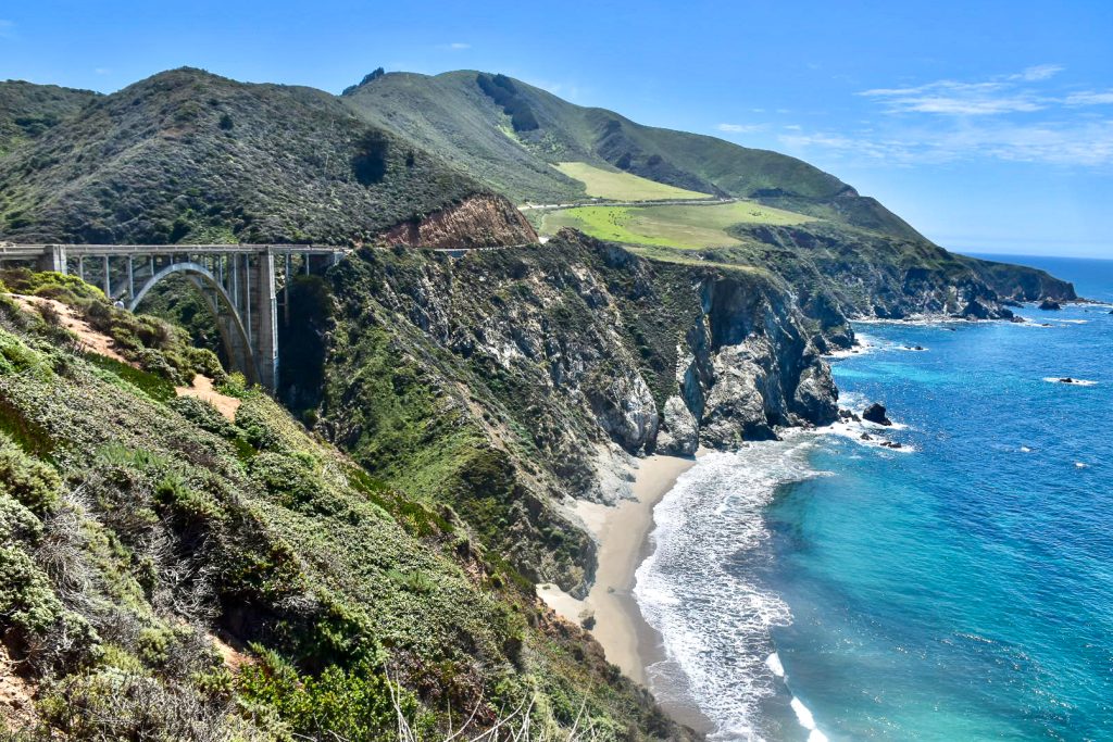 A verdant green hill side with cliffs down to a turquoise and blue sea, runs from the left of the frame across the middle. You can see a single arch road bridge over a ravine and the road running along the cliff side. Pale blue sky above
