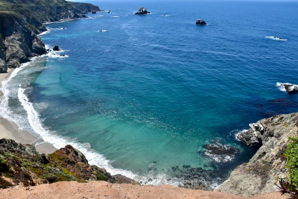 Photo looking down into a cove of flat blue and turquoise waters with small waves at the shore of the crescent bay. You can see the green rocky cliffs around the edge to the left