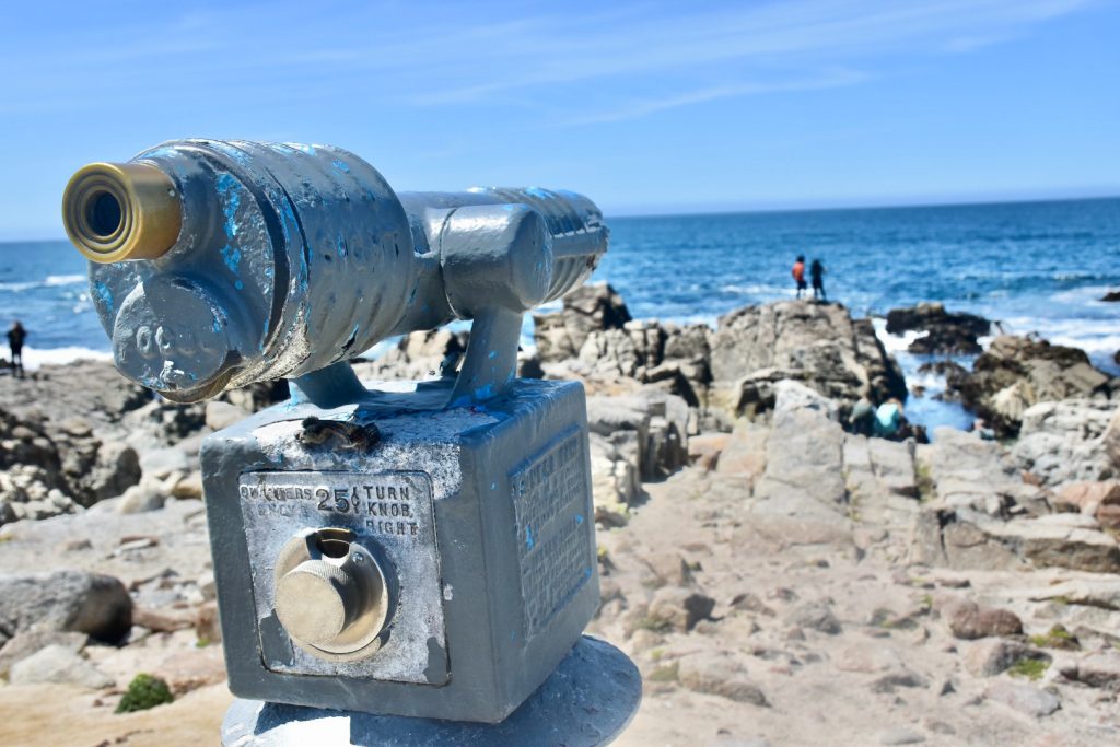 A old-fashioned silver view-finder sits in the bottom left of the photo, looking out across a rocky shoreline with the sea behind and blue sky above