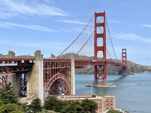 View across a red suspension bridge stretching across a bat. Blue sky. Trees in the foreground