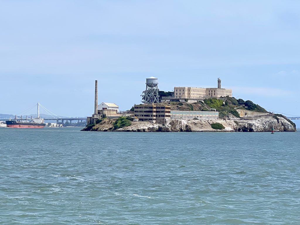 Blue-grey sky at the top, grey-blue sea a the bottom. Across the horizon is an island with prison buildings on it. A bridge and oil tanker in the background on left hand side