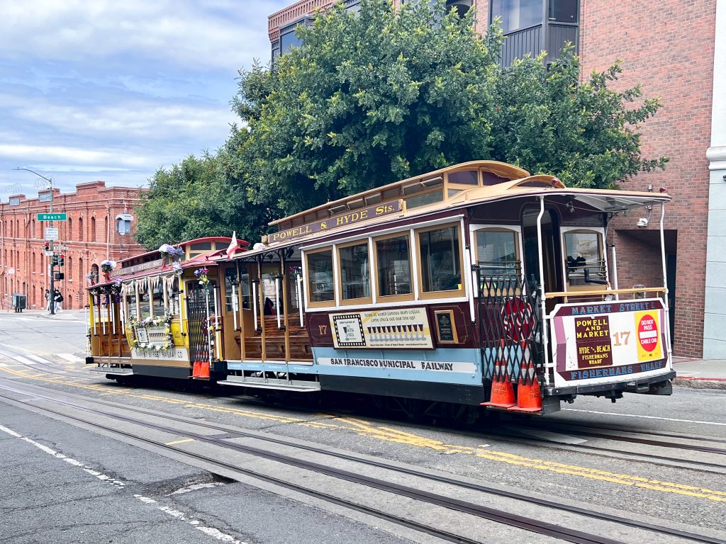 An old fashioned cable car travelling up hill across the front of the photo on tracks. Trees and brick buildings behind. The cable car says Powell & Hyde Street on the top. No passengers