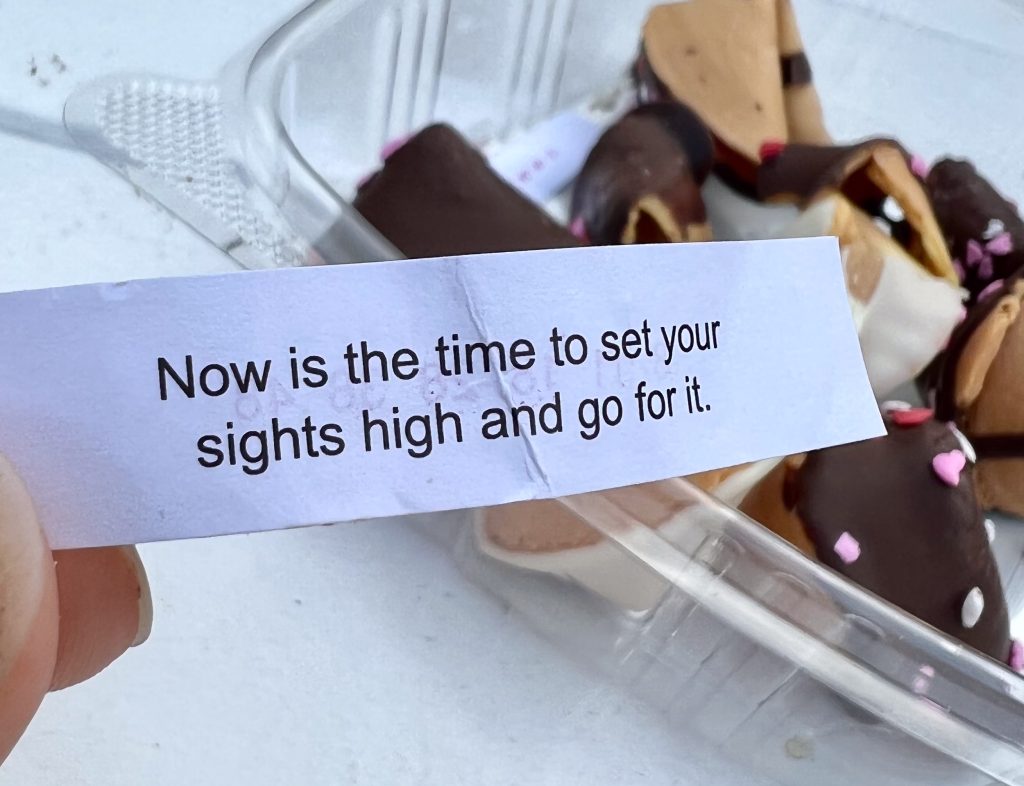 Message from a fortune cookie says 'Now is the time to set your sights high and go for it'. Open container of fortune cookies dipped in chocolate sit on the table behind