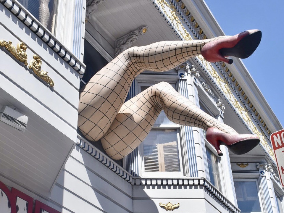 A pair of sculpture legs with tights and red stillettos coming out of the upstairs window of a wooden painted building