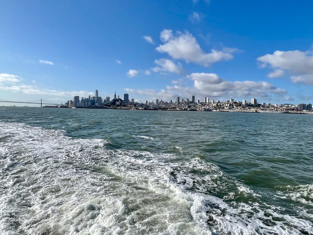 View taken from the back of a boat with the wake visible in bottom left. On the horizon is a cityscape and a long suspension bridge to the left. Blue sky with a handful of white clouds above