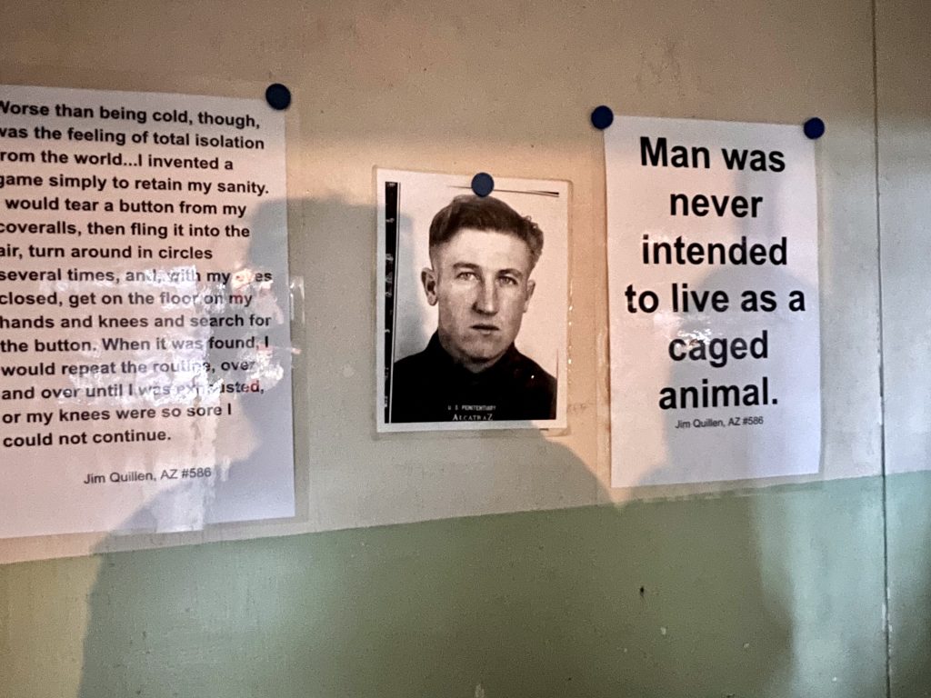 White posters on a concrete wall - poster on the left is a quote about being in solditary confinement by Jim Quillen, the middle photo is a black and white photo of Jim Quillen. Poster on the right says 'Man was never intended to live as a caged animal'.