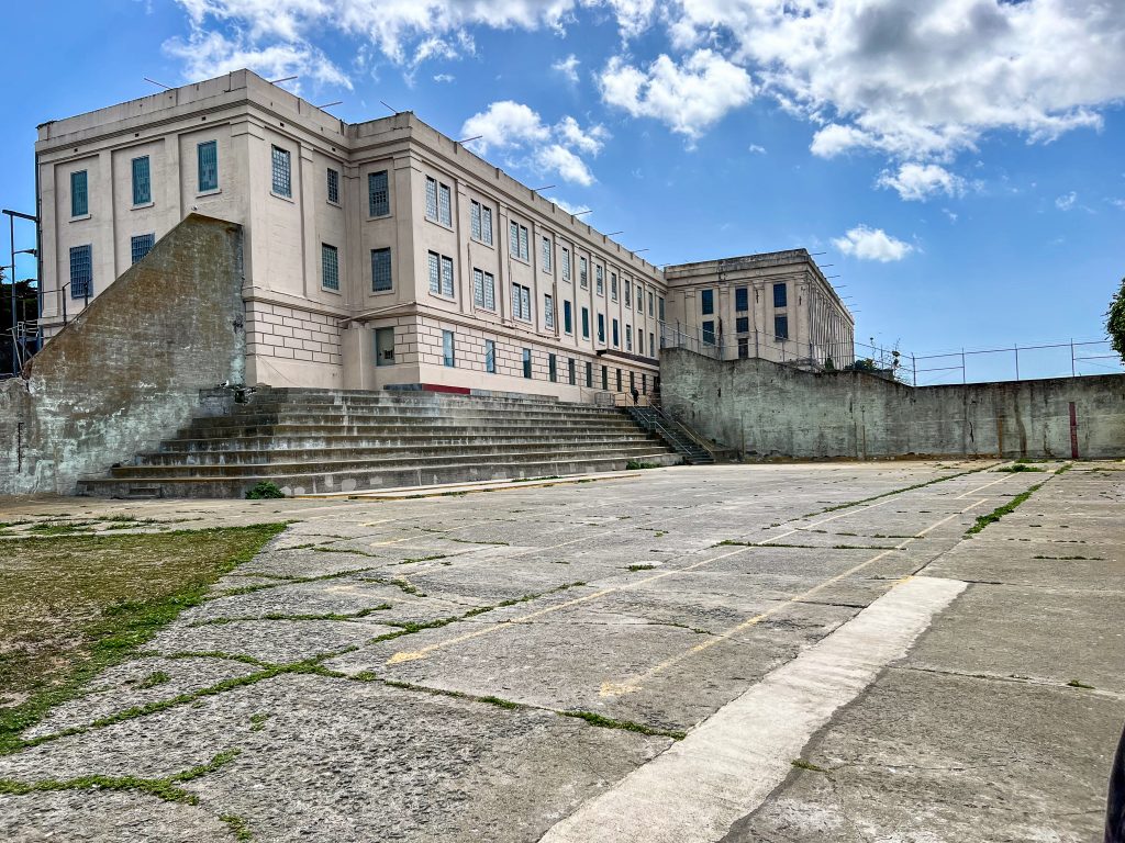 Foreground is a concrete playground, with austere concrete building to the top right hand side. Concrete walls around the edges. Blue sky and white cloud above