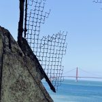 View through cut wire across a bay of blue sea and blue sky with a red suspension bridge in the background