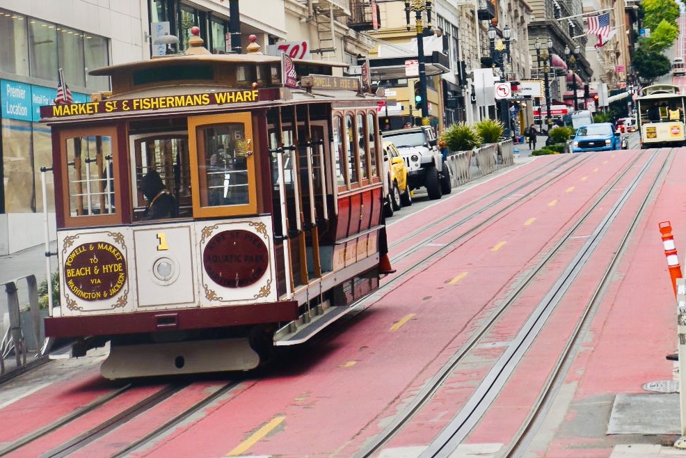 Old fashioned cable car travelling down hill to bottom left of the photo on tracks. Trees and brick buildings behind. A second cable car visible higher up the tracks in the background. Buildings and American flag to the left