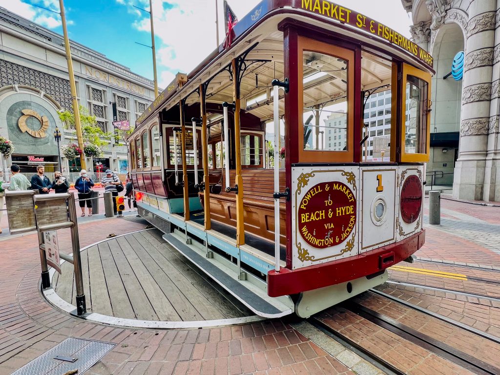 Old fashioned cable car on turning circle with shops in background. Cable car says Market St & Fishermans Wharf on front
