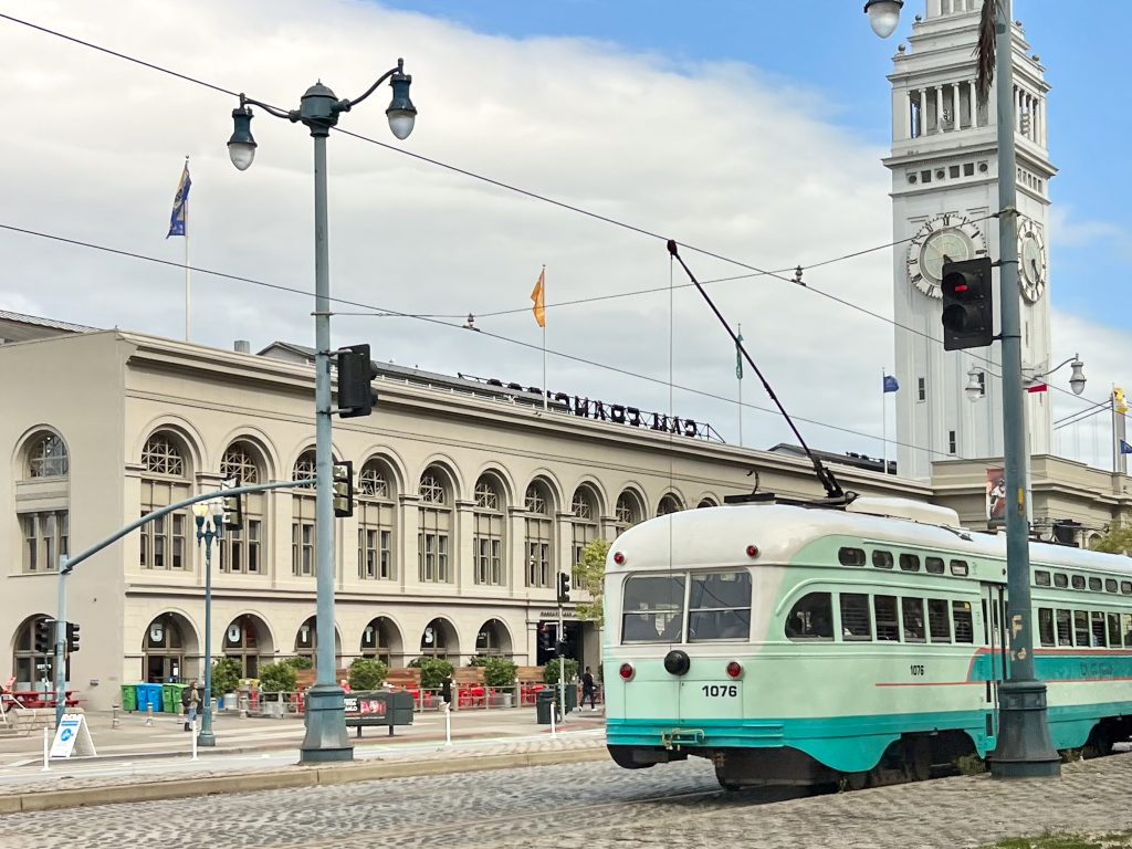 Mint green trolly car enters photo from the bottom right of photo. It is attached to overhead wires by trolley bar. Long white warehouse style building with a white tower in right hand side. Cobble in foreground