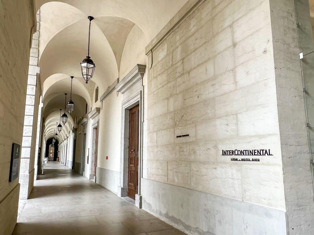 View down an external corridor of white stone with arches and black hanging lanterns all the way down. On the right hand side is a sign saying InterContinental in black letters on white stone brick wall