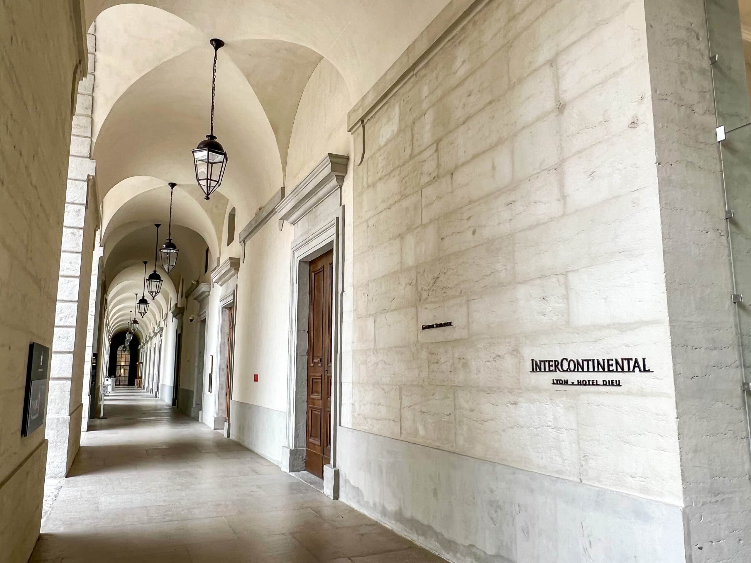 View down an external corridor of white stone with arches and black hanging lanterns all the way down. On the right hand side is a sign saying InterContinental in black letters on white stone brick wall
