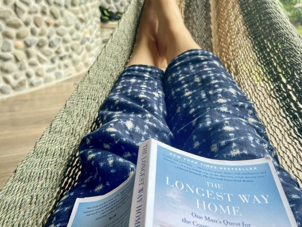 Photo of legs stretched out in a green and yellow knitted hammock. The person is wearing blue and white cotton trousers. A book is open on the legs - facing upward - the book is The Longest Way Home