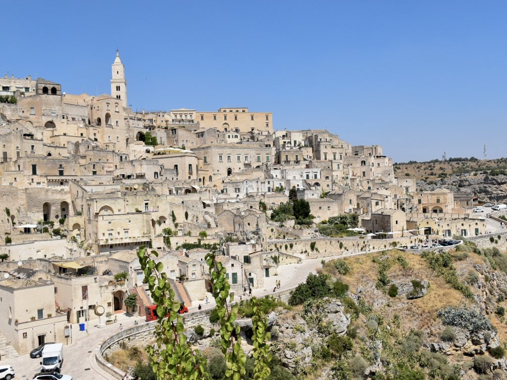 Hillside full of pale coloured stone buildings. A road curves around the bottom of the town at the top of grass and rock cliff. Blue sky above
