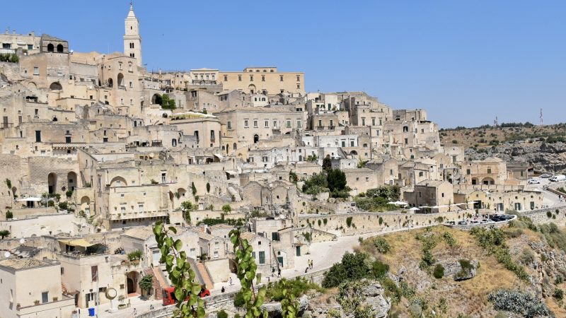 Hillside full of pale coloured stone buildings. A road curves around the bottom of the town at the top of grass and rock cliff. Blue sky above