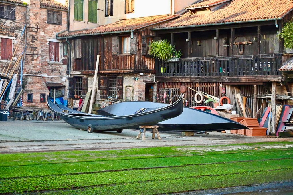 Two black gondolas site outside wooden workshop builings - one sits on a bench. There is grass on the ramp in front leading to the water