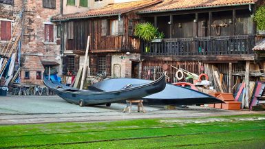 Two black gondolas site outside wooden workshop builings - one sits on a bench. There is grass on the ramp in front leading to the water