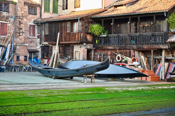 Two black gondolas site outside wooden workshop builings - one sits on a bench. There is grass on the ramp in front leading to the water
