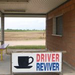 Image taken out of a car window. An outdoor patio area with corrugated metal roof and red brick walls to the right. Bench to left. A white sign says 'Driver (in red) Reviver (in blue)' with a black cup of tea image, at the bottom of the shot