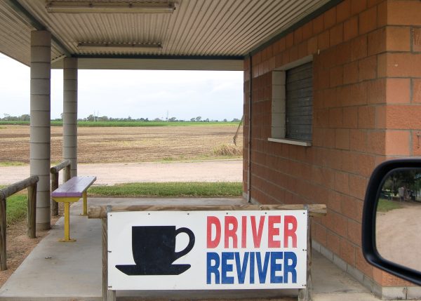 Image taken out of a car window. An outdoor patio area with corrugated metal roof and red brick walls to the right. Bench to left. A white sign says 'Driver (in red) Reviver (in blue)' with a black cup of tea image, at the bottom of the shot