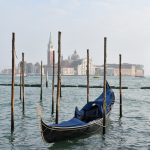 Bottom half features waters of a blue canal, a gondola parked facing the camera and wooden posts sticking out of the water. There are buildings on the horizon and a campanile. Grey sky behind