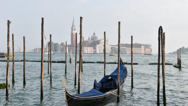 Bottom half features waters of a blue canal, a gondola parked facing the camera and wooden posts sticking out of the water. There are buildings on the horizon and a campanile. Grey sky behind