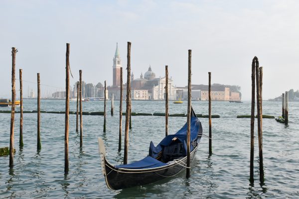 Bottom half features waters of a blue canal, a gondola parked facing the camera and wooden posts sticking out of the water. There are buildings on the horizon and a campanile. Grey sky behind