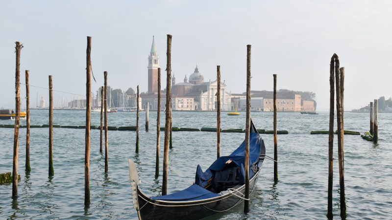 Bottom half features waters of a blue canal, a gondola parked facing the camera and wooden posts sticking out of the water. There are buildings on the horizon and a campanile. Grey sky behind