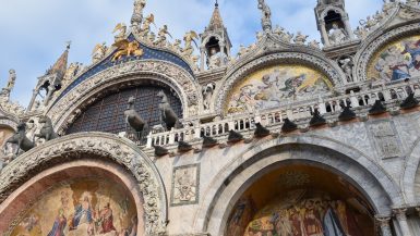 Upwards photo of a particularly ornate building, looks like a church. Arches with mosaics and paintings. Balcony with horse statues. Statues on top