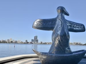 Silver statue of a bird with outstretched wings sitting in a boat shape - indigenous art design. Overlooks a river with city buildings on the horizon. Pale blue sky above