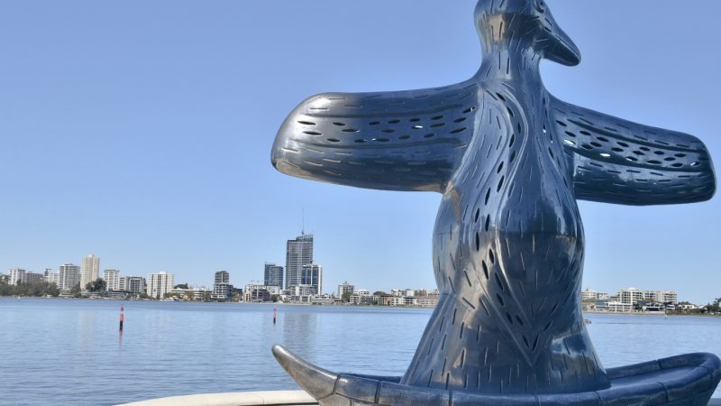 Silver statue of a bird with outstretched wings sitting in a boat shape - indigenous art design. Overlooks a river with city buildings on the horizon. Pale blue sky above