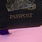 Black leather Passport holder with a golden emblem printed on the front and the word Passport. Purple ribbon popping out of the bottom. On a sugar pink lined paper