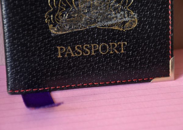 Black leather Passport holder with a golden emblem printed on the front and the word Passport. Purple ribbon popping out of the bottom. On a sugar pink lined paper